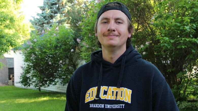 A young man wearing a cap and a Brandon University faculty of education sweatshirt smiles as he stands in a garden. 