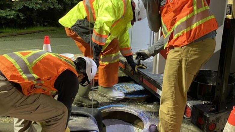 Men in safety vests hunch over around a large hole in the ground. 