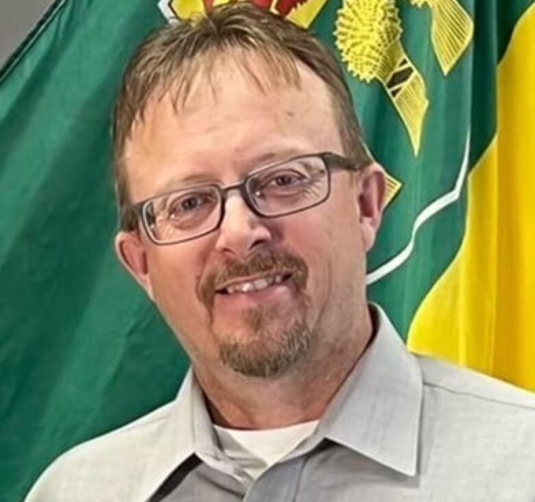 A man with classes stands in front of a furled Saskatchewan flag.