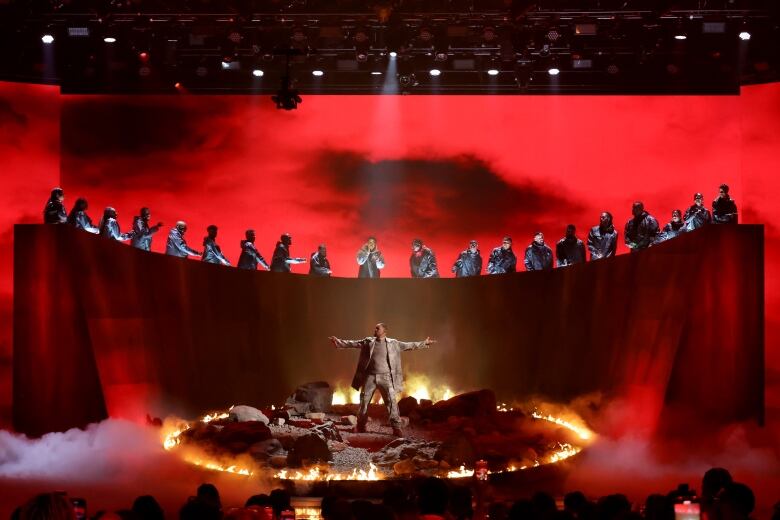 Will Smith, his arms outstretched, stands in a circle of fire on stage. Other performers stand above him on a raised, curved wall. Above it all stage lights are visible.