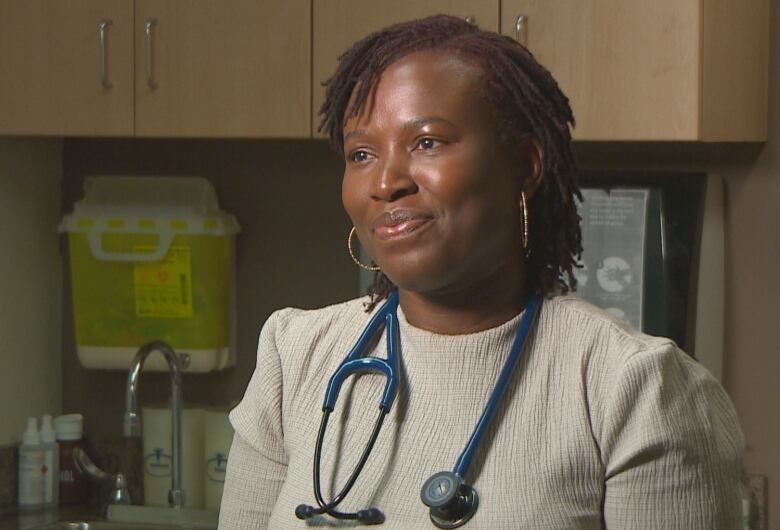 A woman with a stethoscope around her shoulders stands in an exam room at a doctor's office.