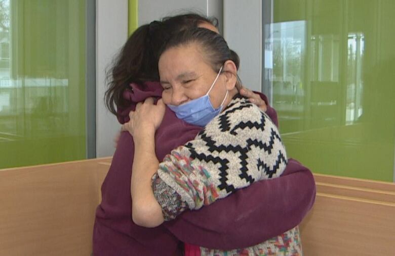 A woman in a checked shirt holds another younger woman in a purple hoodie in her arms.