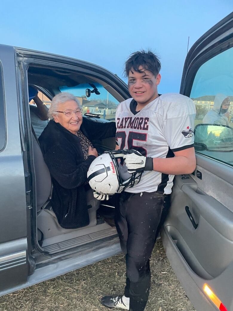 A white-haired woman in a truck sits on the passenger side while beside her stands a young man in a white football jersey, holding a helmet.