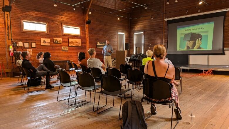 A group of people in chairs looks at an image on a screen in wood panelled room. 