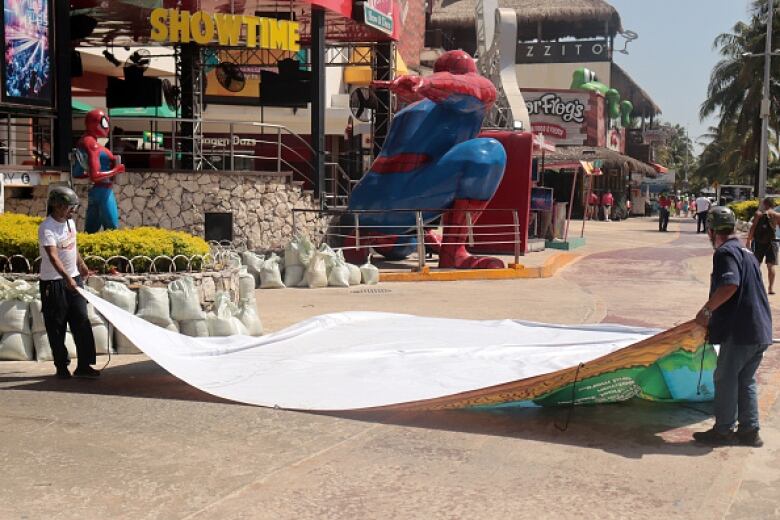 Two men wearing helmets hold a large tarp on a road at a resort community.