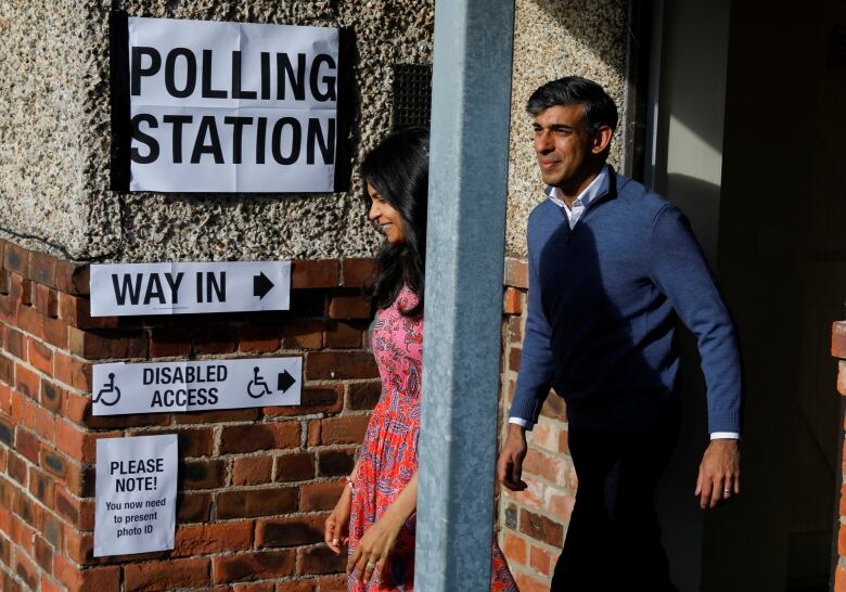 A man and woman walk out of a building plastered with signs that read 'Polling Station' and 'Way In,' among other things.