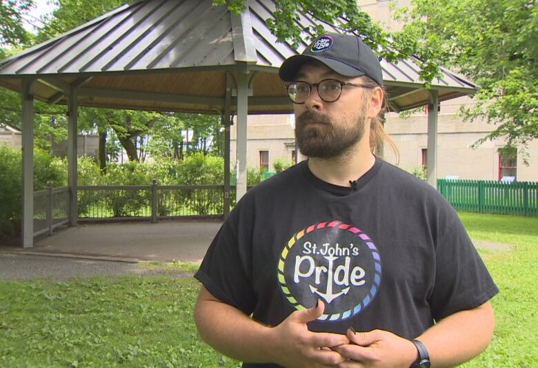 A bearded man with glasses and a baseball cap is in a park. His T-shirt has a rainbow-coloured circle with the words 'St. John's Pride' inside it.