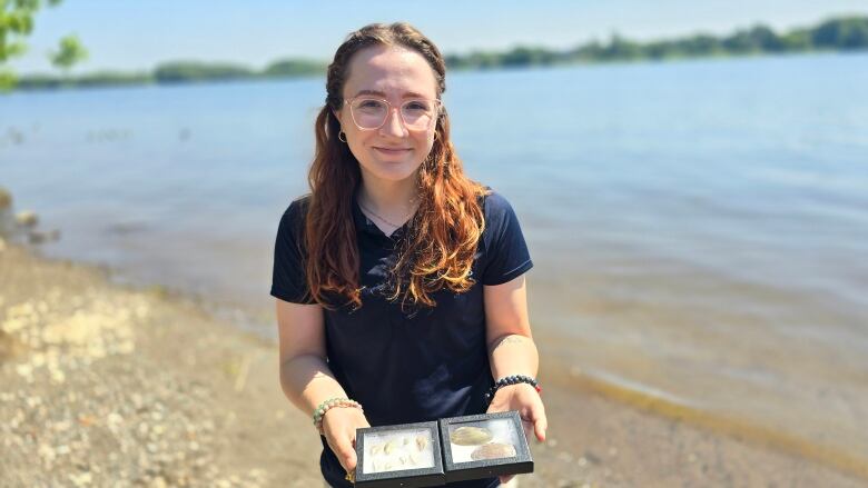 Woman stands in the sun on the shore of a river holding display cases