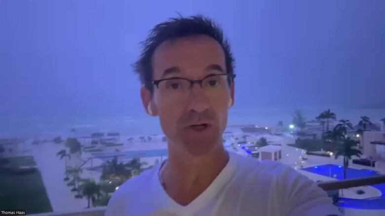 A man in glasses in front of a pool and palm trees, as lightning strikes in the background. 