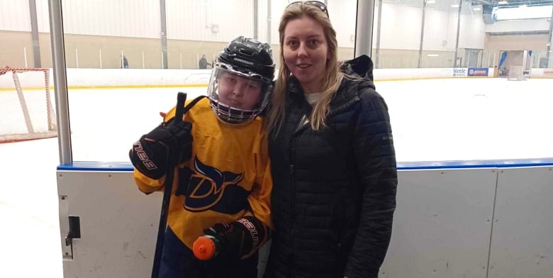 A young boy wears black and yellow hockey gear. He is posing with his mother in front of a hockey rink. 