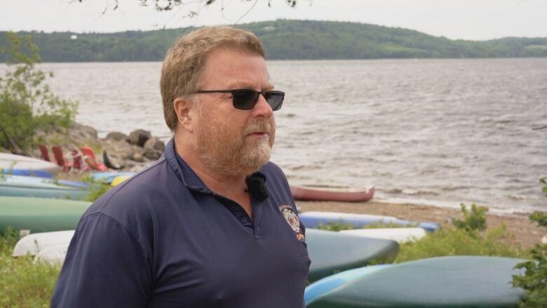 Man in blue shirt and sunglasses standing on the edge of a beach with a body of water behind him. 