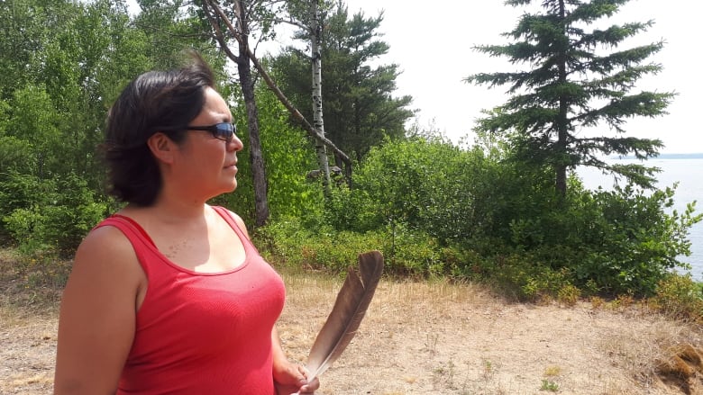 A person wearing a bright pink tank top and sunglasses is seen holding an eagle feather while standing on the beach. Behind them are green trees. They are looking toward the water.