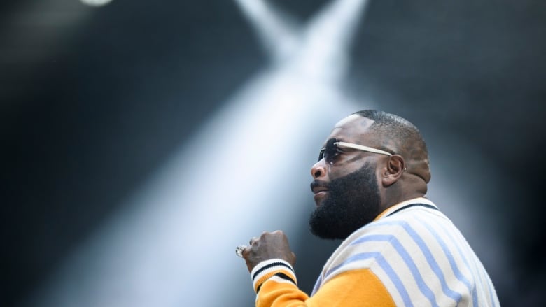 A middle-aged Black man stands on a stage wearing a blue and yellow striped jacket and black sunglasses.