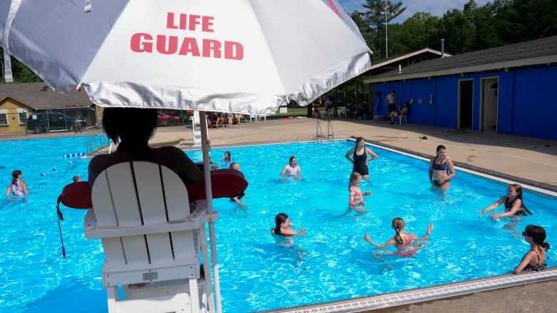 A lifeguard sitting in a high chair watches kids swim in a pool.