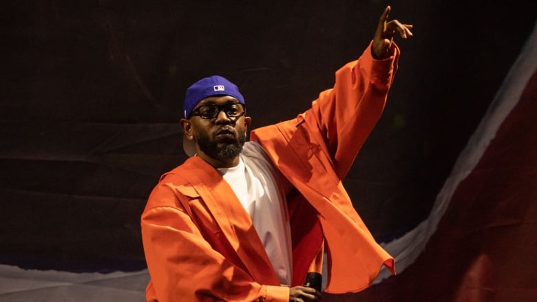 A man in an orange athletic suit and purple ball cap holds a microphone in one hand and gestures to the sky with another as he performs on stage.