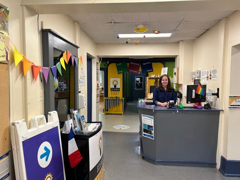 An entryway with a blue counter, blue directional arrow sign, rainbow flags, a solar system model hanging from the ceiling, and the Science East yellow and black logo with a lightbulb on the wall in the background.