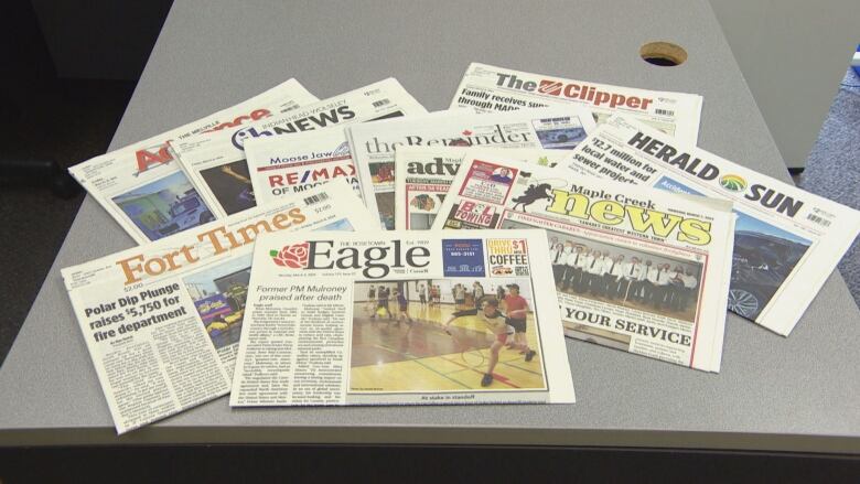 Numerous weekly newspapers from Saskatchewan are spread out on a table.