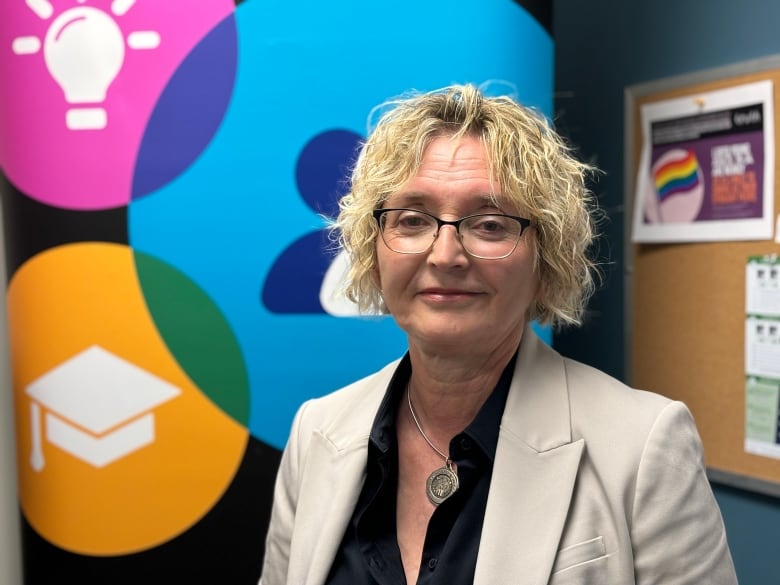 A woman wearing a black blouse and white blazer stands in front of a colourful sign.