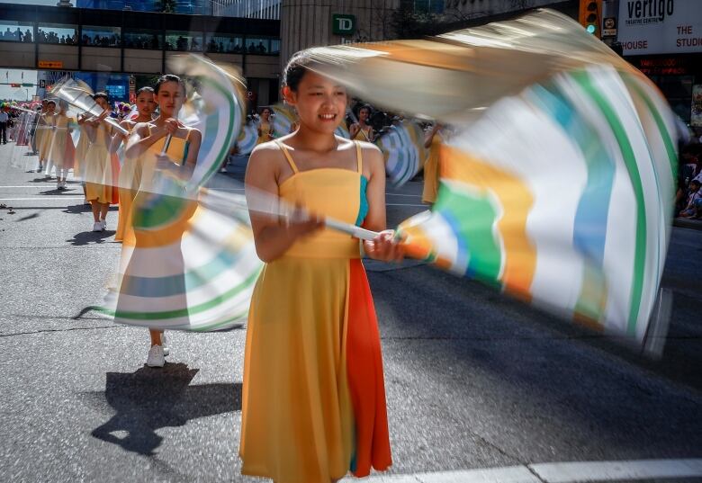 girls in yellow dresses walk in a parade twirling sticks with flowing fabric.