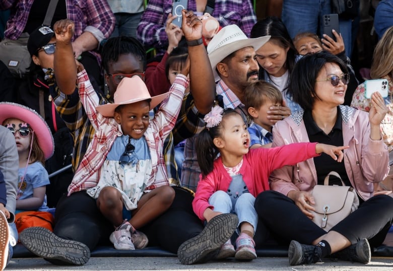 Children and parents sit on a curb while watching the Stampede. One child is smiling with hands in the air. Another is pointing at something she sees.