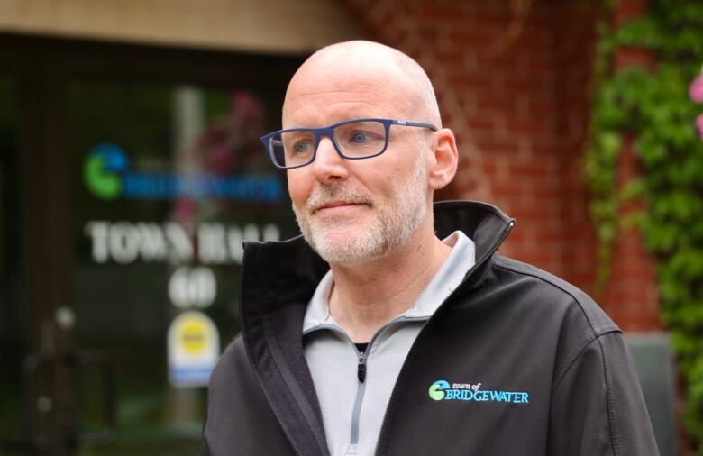 Bald man with glasses and beard wears black jacket with 'Town of Bridgetown' label. 