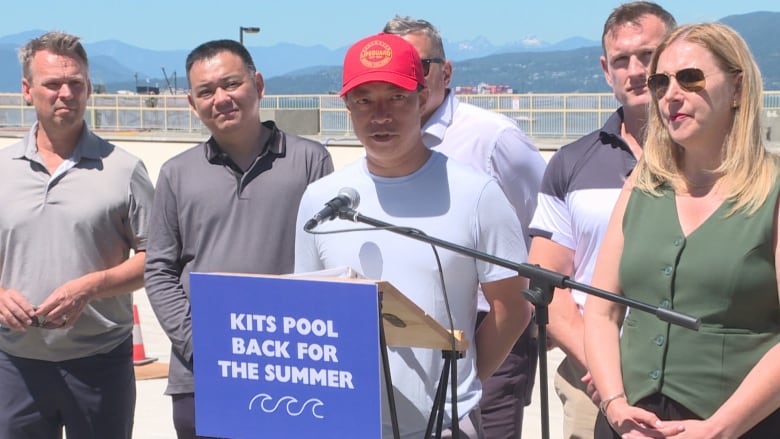 Politicians stand at a microphone with an empty pool in the background.