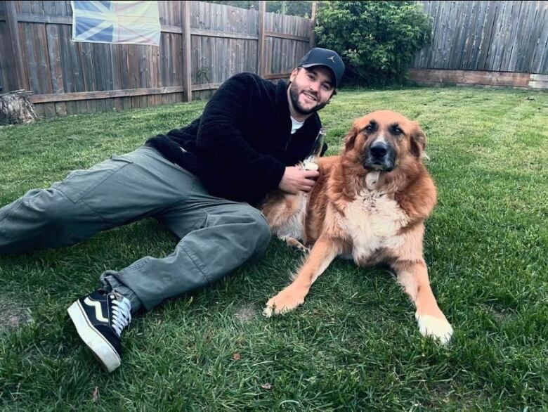 A man and a golden retriever lay in fresh cut grass. In the background, a large Newfoundland flag is visible.