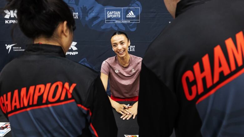 A woman sits behind a table and smiles to two people standing in front of her.