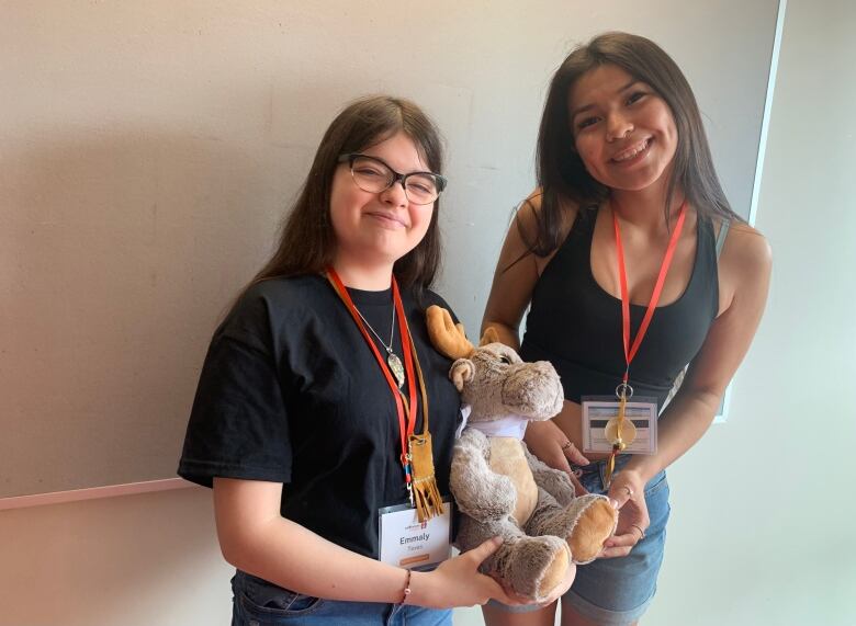 Two students stand in front of a white wall holding a stuffed moose.