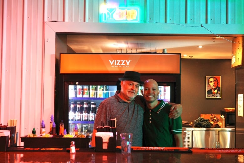 A gay couple stand behind a bar inside a nightclub. Behind them is a white wall, which is pink and blue from nearby fluorescent lighings. The man on the left is older and wears a fedora and a grey golf shirt. The man on the right is bald and wears a polo shirt. 