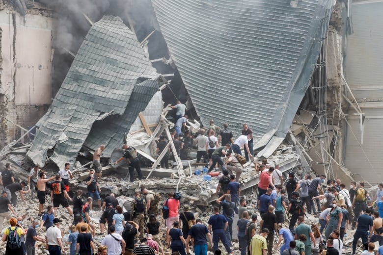 Dozens of people are shown at a distance standing on rubble near a building with a heavily damaged section.