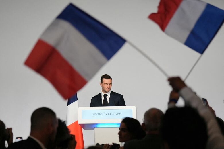 A young man in a suit stands at a podium with a crowd cheering around him.