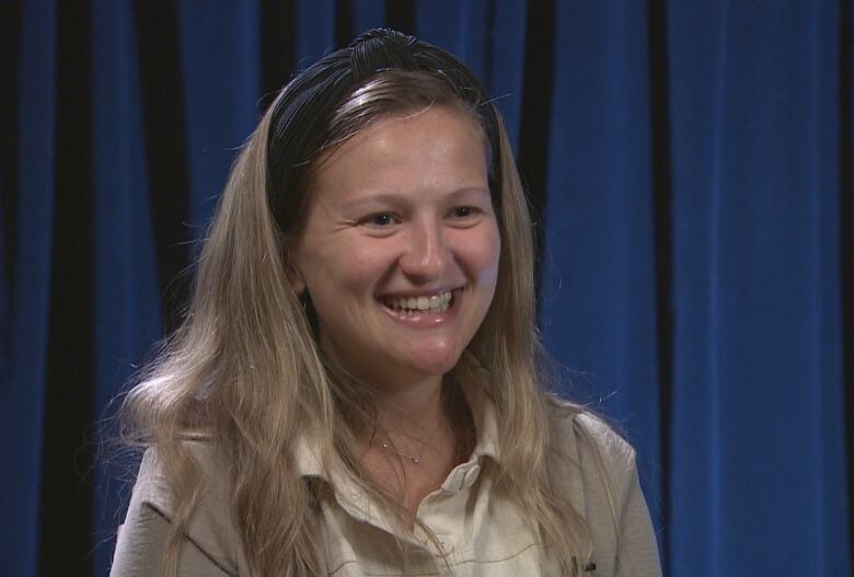 A smiling woman wears bandana on her head. She is seated before a blue curtain. 