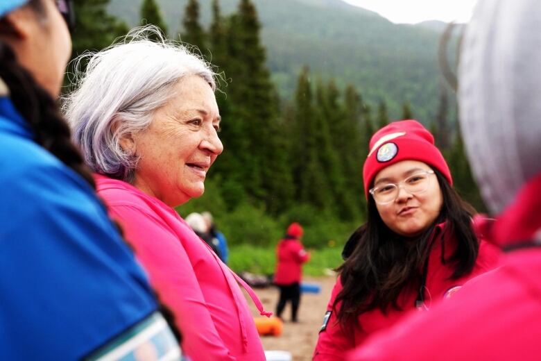 A woman wearing a pink sweater talks to teenagers wearing red and blue. 