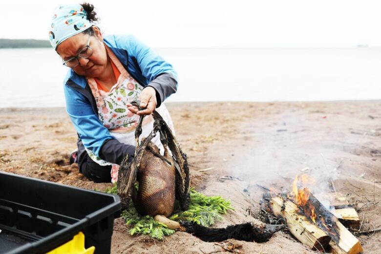 A woman holds a goose without feathers by a firepit in sand. 