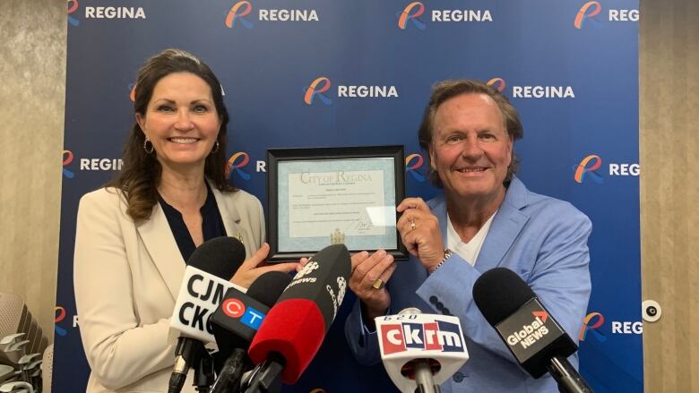 Two people in suits hold up a framed certificate in front of media microphones. 