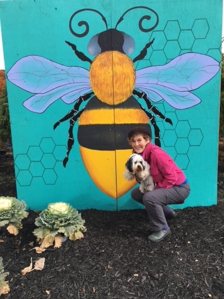 A woman in a purple jacket holding a small white dog crouching in front of a sign of a painted honey bee