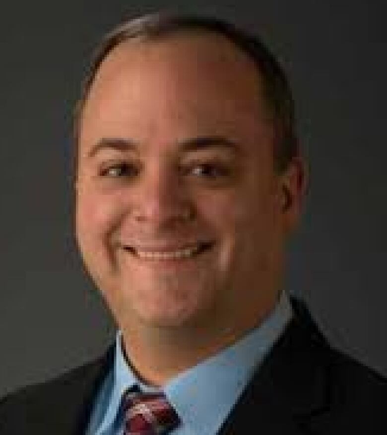 Dr. Lloyd Mack is smiling and wearing a black suit, a red plaid tie and blue dress shirt.