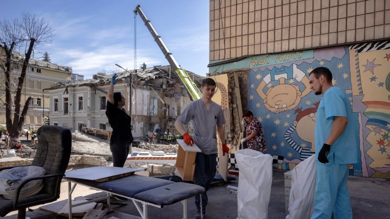 A man in medical scrubs and other people carry items outside a heavily damaged building.