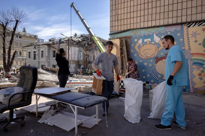 A man in medical scrubs and other people carry items outside a heavily damaged building.