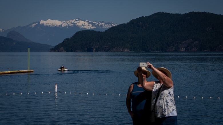 Two people in hats take a selfie in front of a lake. A personal watercraft can be seen on the lake, and snow-capped mountains in the distance.