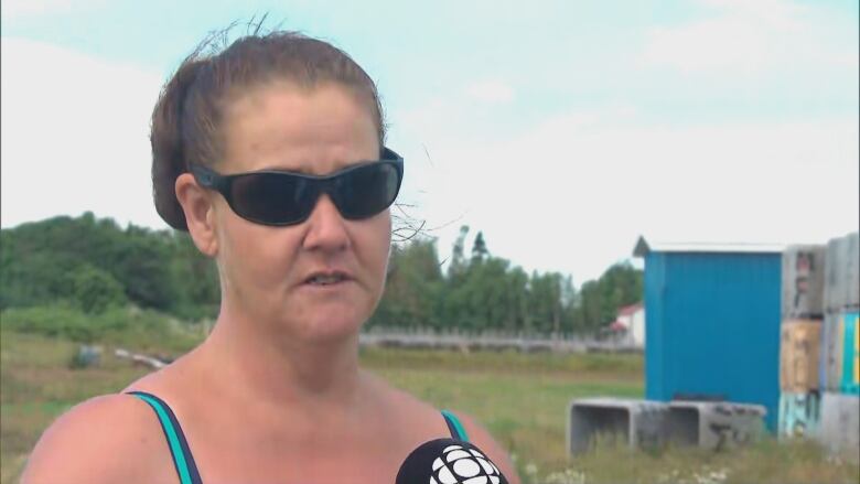 A woman with her brown hair in a pony tail and wearing glasses and a tank top faces the camera in a head and shoulders shot. the top of a microphone with the CBC logo can be seen at the bottom of the frame below her mouth. In the background are some blue and gray fishing bins, a grassy field and a line of leafy trees against a white sky.