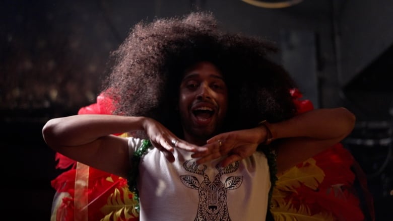 A person wearing bright red and yellow feathers and a big Afro wig dances on a stage.