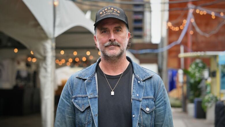 A man stands in the foreground with a baseball cap on and a jean jacket, behind him a tent and lights are visible.