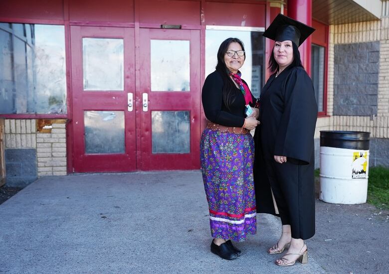 Two Innu women stand in front of a school. One is wearing a graduation gown and cap, while the other wears a black sweater and purple ribbon skirt. 