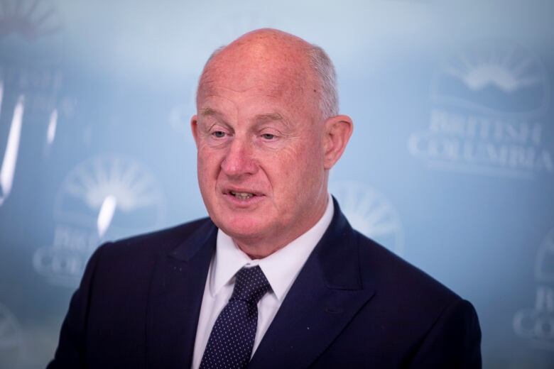 A balding white man wearing a suit speaks in front of a wall with indented 'British Columbia' logos.