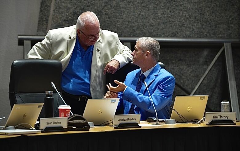 man in white suit jacket leans on a chair speaking to a seated man in a blue suit