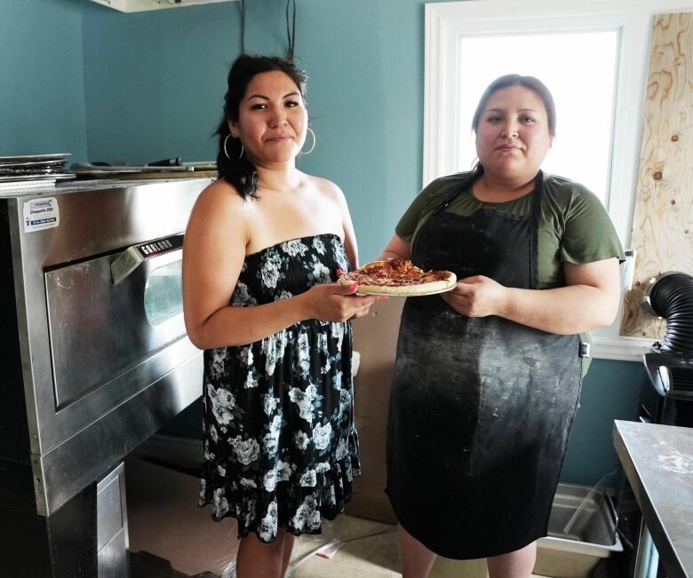 Two women stand in front of a pizza oven, holding a pizza. 