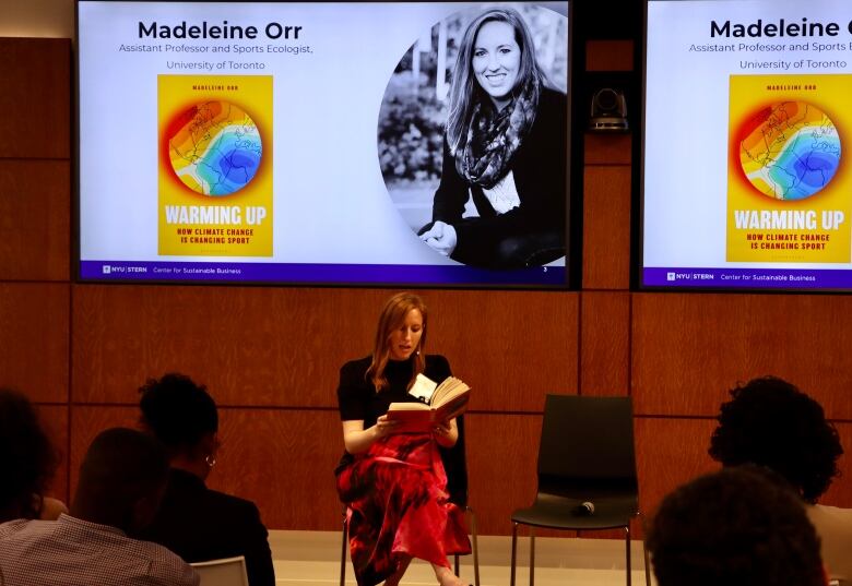 A woman reads to an audience from a book, in front of a screen showing her name Madeleine Orr and book title Warming Up.