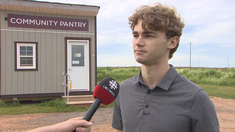 A man in a grey shirt with dark blond curly hair stands in front of a shed labelled Community Pantry.